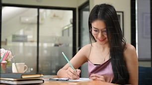 Asian woman is writing on a clipboard while sitting at the wooden working desk.