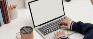 Ausschnittaufnahme einer Freiberuflerin, die auf einem Mock-up-Laptop auf einem weißen Schreibtisch mit Eiskaffeetasse, Büchern und Schreibwaren im Home Office tippt
