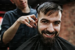 Good looking young adult man getting a hair and beard styling and dressing treatment by professional hairstylist.