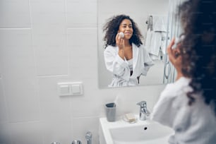 Cute joyous woman in a bathrobe performing a beauty procedure in front of the mirror
