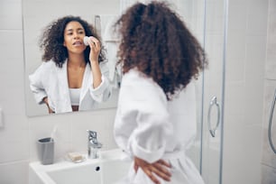 Fokussierte junge Frau mit einer doppelseitigen Gesichtsbürste aus Silikon, die vor dem Spiegel steht