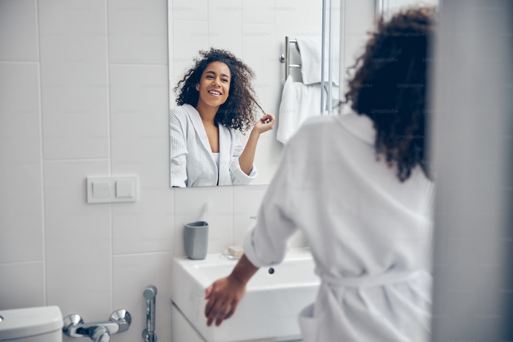 Mulher alegre brincando com uma mecha de seu cabelo encaracolado na frente do espelho