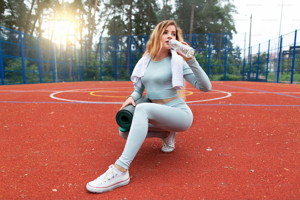 灰色のスポーツウェアを着て、ボトルから水を飲み、ヨガのスポーツマットを持ち、屋外の赤い床のスポーツスタジアムでスポーツエクササイズの後にリラックスしている若い白人女性。健康的なライフスタイル。