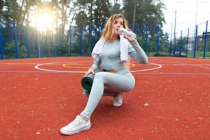 Young caucasian woman wearing gray sportswear, drinking water from bottle, holding a yoga sport mat and relaxing after sport exercises on sport stadium with red floor, outdoor. Healthy lifestyle.