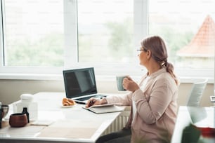 Senior woman working from home during the quarantine looking at the window while drinking tea with croissant and work at computer
