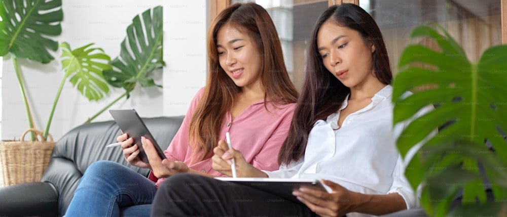 Cropped shot of female teenagers relaxed sitting on black couch, pink shirt woman using smartphone while white shirt woman using tablet on her lap