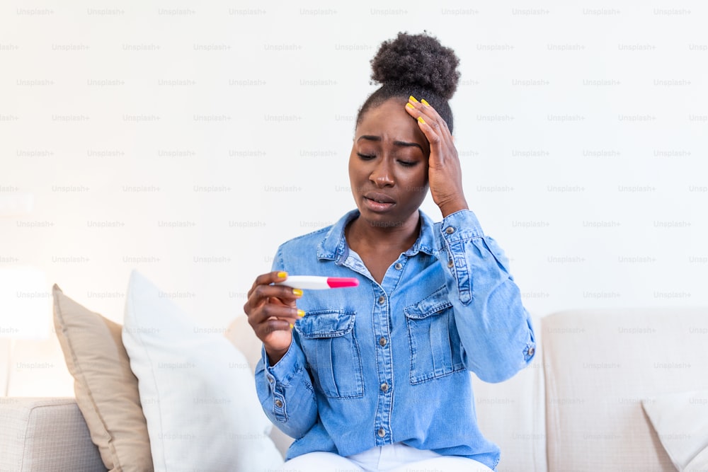 Single sad woman complaining holding a pregnancy test sitting on a couch in the living room at home. Depressed Black girl holding negative pregnancy test.