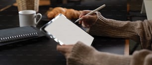 Side view of female hands using mock up tablet while sitting at breakfast table with schedule books, coffee and croissant