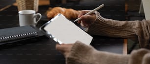 Vista lateral de las manos femeninas usando una tableta de maqueta mientras está sentada en la mesa del desayuno con libros de horarios, café y croissant