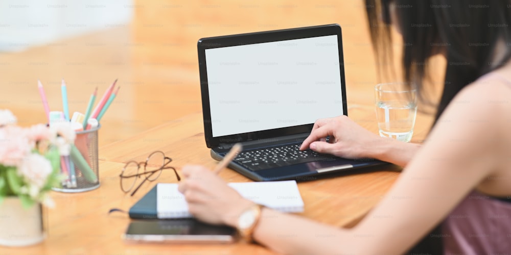 A mulher de imagem cortada está usando um laptop de computador de tela branca em branco na mesa de trabalho de madeira.