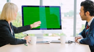 オフィスのテーブルの上にグリーンスクリーンのクロマキーテレビやコンピューターがある会議室のビジネスマン。ビデオ会議通話で会議中のビジネスマンとビジネスウーマンの多様なグループ。