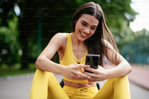 Beautiful woman resting from working out. Young athlete woman sitting on bench and using the phone.