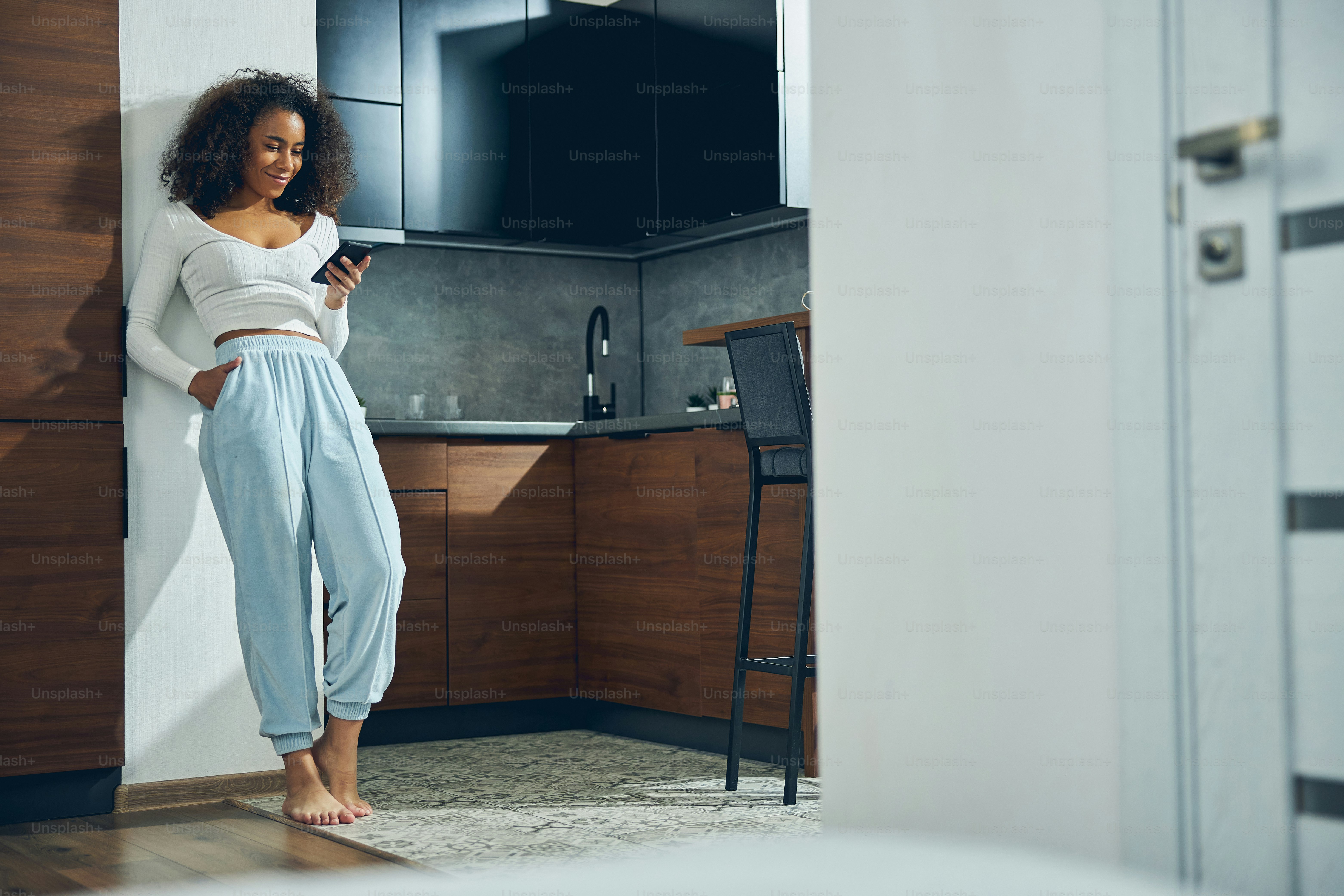Beautiful African American female leaning against the wall in the kitchen while holding her mobile and smiling