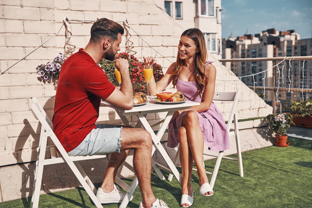 Cute young couple in casual clothing having breakfast and smiling while sitting on the rooftop patio outdoors