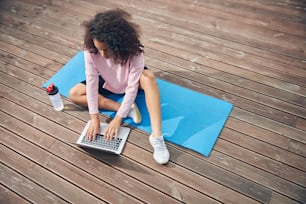 African American female in sports outfit sitting on special yoga mat and spending time with laptop