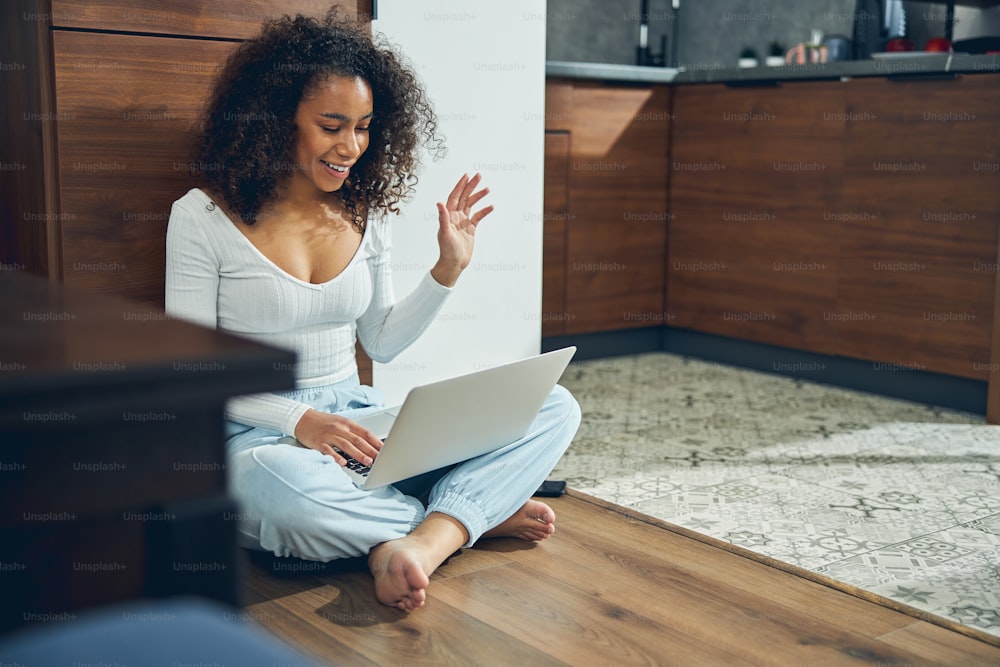 Cheerful woman sitting with her legs crossed in front of the laptop on the kitchen floor