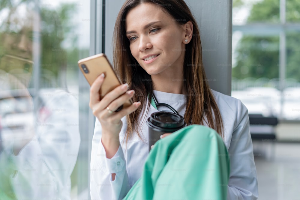 白衣を着た若い女性医師の肖像画は、病院でスマートフォンを使用しながら座り、仕事の後にリラックスします。