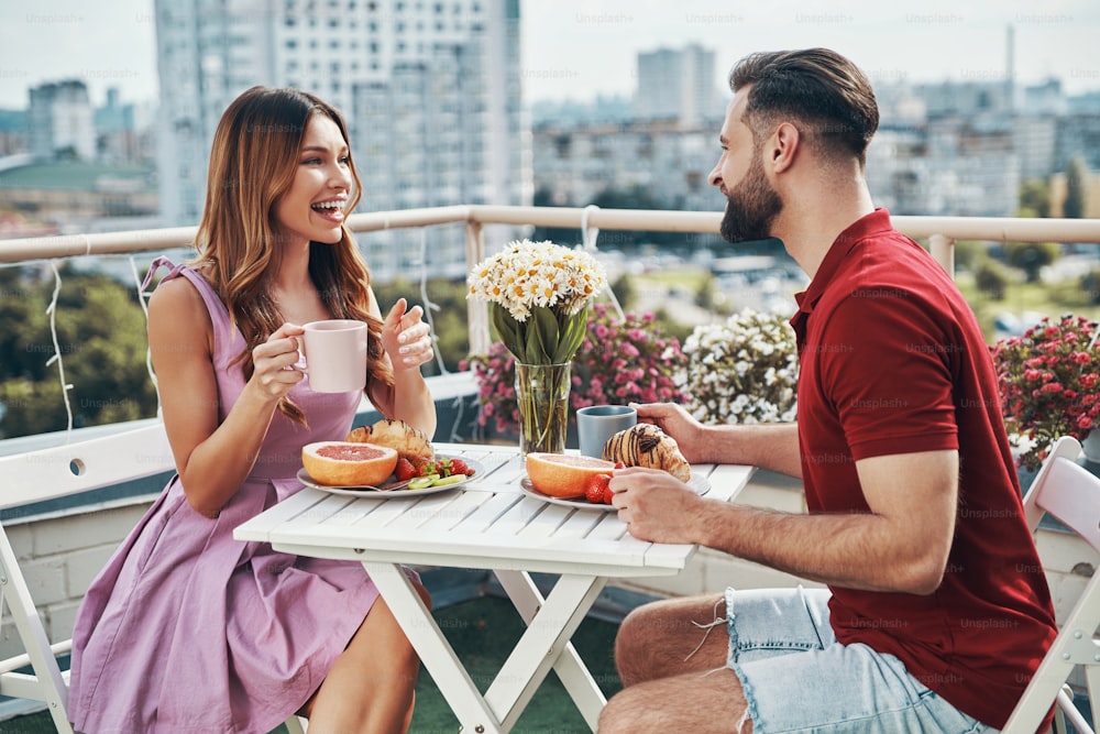 Glückliches junges Paar in Freizeitkleidung, das Abendessen genießt und lächelt, während es auf der Dachterrasse im Freien sitzt