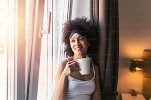 Retrato de una mujer negra sonriente, feliz, alegre, hermosa, bonita, relajada, bebiendo, tomando una taza de café o té caliente y mirando por la ventana. Mujer joven con máscara para dormir tomando café por la mañana