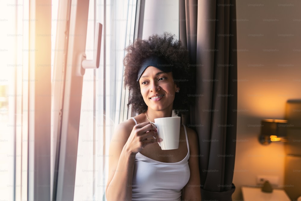 Portrait of smiling happy cheerful beautiful pretty black woman relaxing drinking cup of hot coffee or tea and looking out the window. Young woman with sleeping mask having coffee in the morning