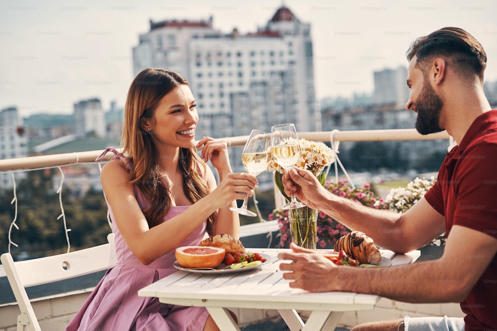 Encantadora pareja joven con ropa informal brindando y sonriendo mientras están sentados en el patio de la azotea al aire libre