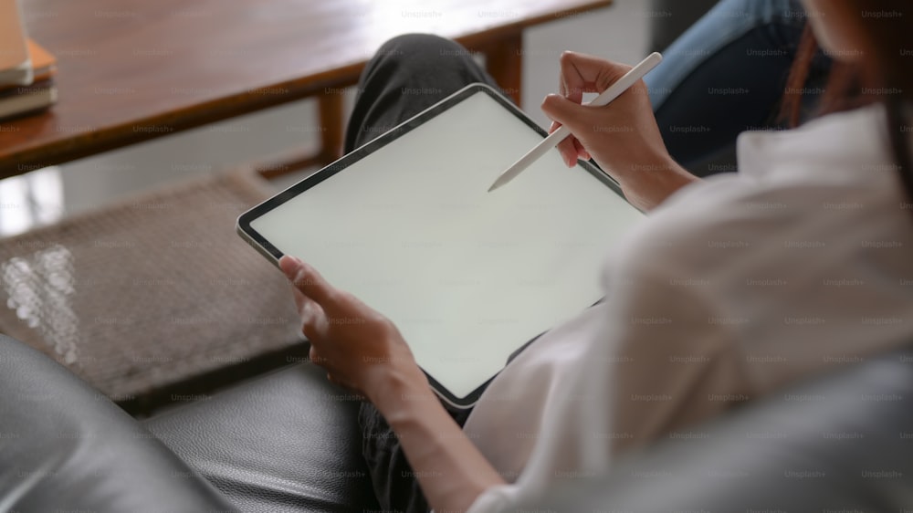 Cropped shot of female university student using mock-up tablet on her lap while relaxed sitting on black couch