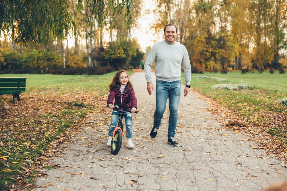 Pai feliz e sua filha andando no parque com uma bicicleta e sorrir para a câmera