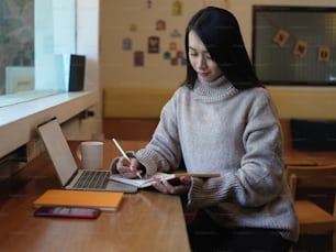 Retrato da estudante universitária feminina que faz o dever de casa com papelaria e laptop no balcão do bar no café