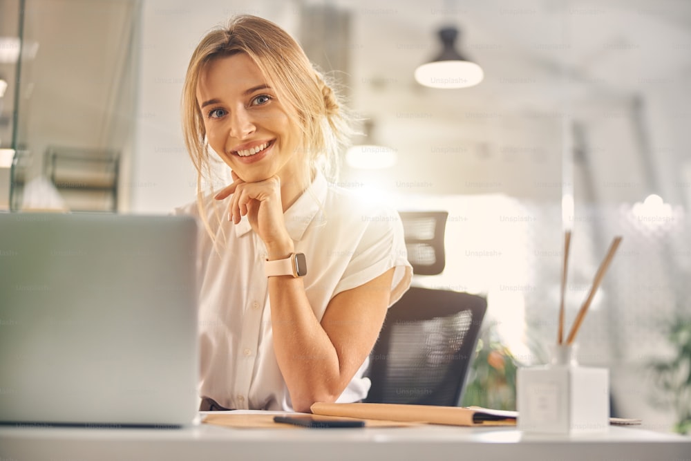 Charming blonde lady looking at camera and smiling while sitting at the table with laptop at work