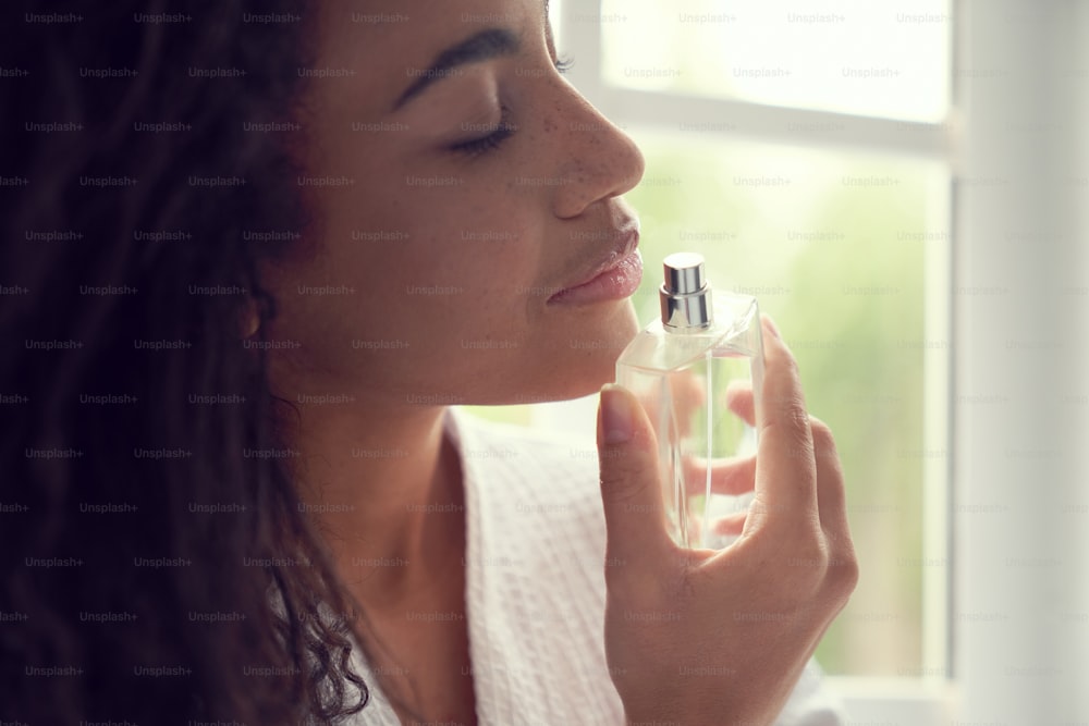 Primo piano di bella giovane femmina riccia che sente l'odore dell'aroma mentre tiene la bottiglia con il profumo in mano