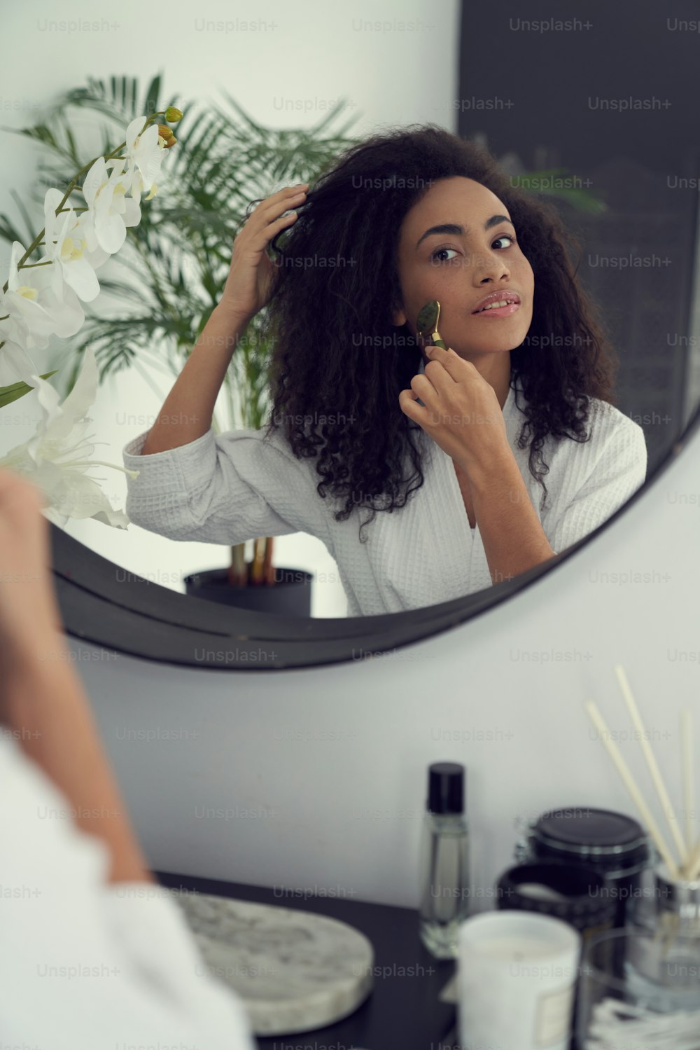 Relaxed curly female is standing in front of mirror in bathroom and using jade roller on cheeks