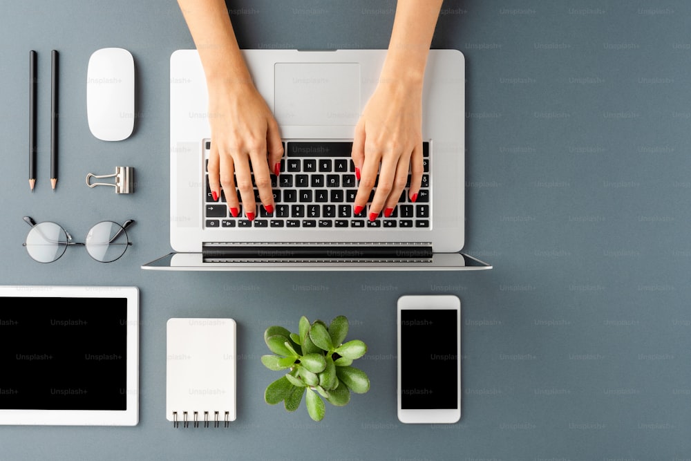 Colpo dall'alto delle mani della donna che lavorano sul laptop sul tavolo grigio con accessori. Desktop dell'ufficio. Posa piatta