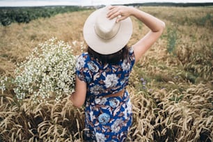 田舎の静かな夏。青いビンテージドレスと帽子をかぶったスタイリッシュな若い女性が、小麦畑の牧草地で白い野の花と一緒にポーズをとっています。大きなヒナギクの花束を持つ美しい女の子、後ろ姿。