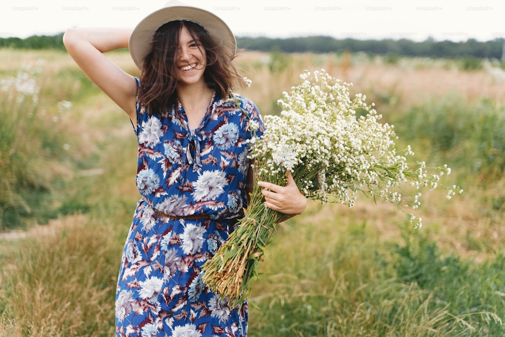 Beautiful girl having fun and laughing in windy field with big daisies bouquet. Authentic summer in countryside. Young woman in blue vintage dress and hat dancing with white wildflowers in meadow.