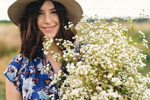 Hermosa mujer joven con vestido vintage azul y sombrero sosteniendo flores silvestres blancas en el prado. Retrato sensual de muchacha hermosa con ramo grande de margaritas en campo ventoso. Verano en el campo