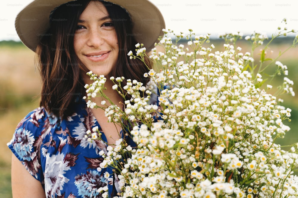 Beautiful young woman in blue vintage dress and hat holding white wildflowers in meadow. Sensual portrait of beautiful girl with big daisies bouquet in windy field. Summer in countryside
