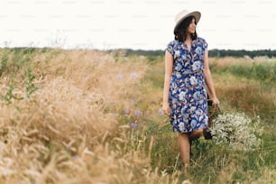 田舎の静かな夏。青いビンテージドレスと帽子をかぶったスタイリッシュな若い女性が、黄色い小麦畑で藁かごに入った白い野の花と一緒に歩いています。白いヒナギクの花束を持つ美しい女の子。