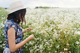 夏の牧草地で白い野の花を集めている青いヴィンテージドレスと帽子を着たスタイリッシュな若い女性。田舎の静かな夏。白いヒナギクに立っている美しい幸せな女の子