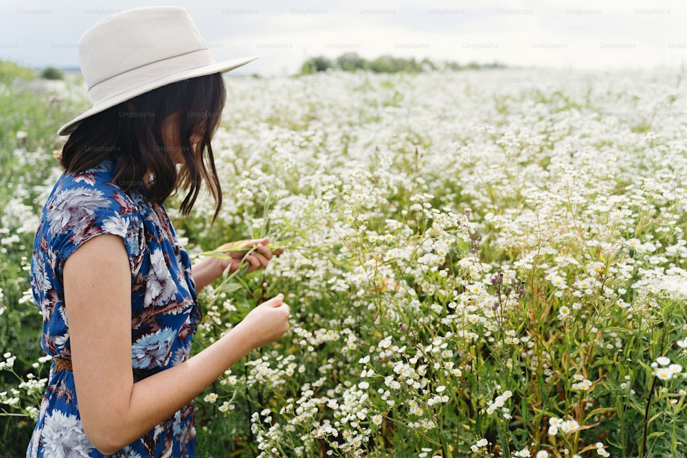Mujer joven con estilo en vestido vintage azul y sombrero que recoge flores silvestres blancas en el prado de verano. Verano tranquilo en el campo. Hermosa muchacha feliz de pie en margaritas blancas