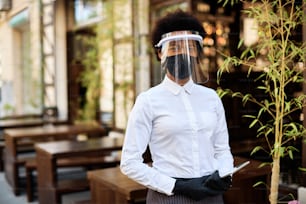 Camarera afroamericana con máscara protectora y visera mientras está de pie en un café al aire libre y mira a la cámara.