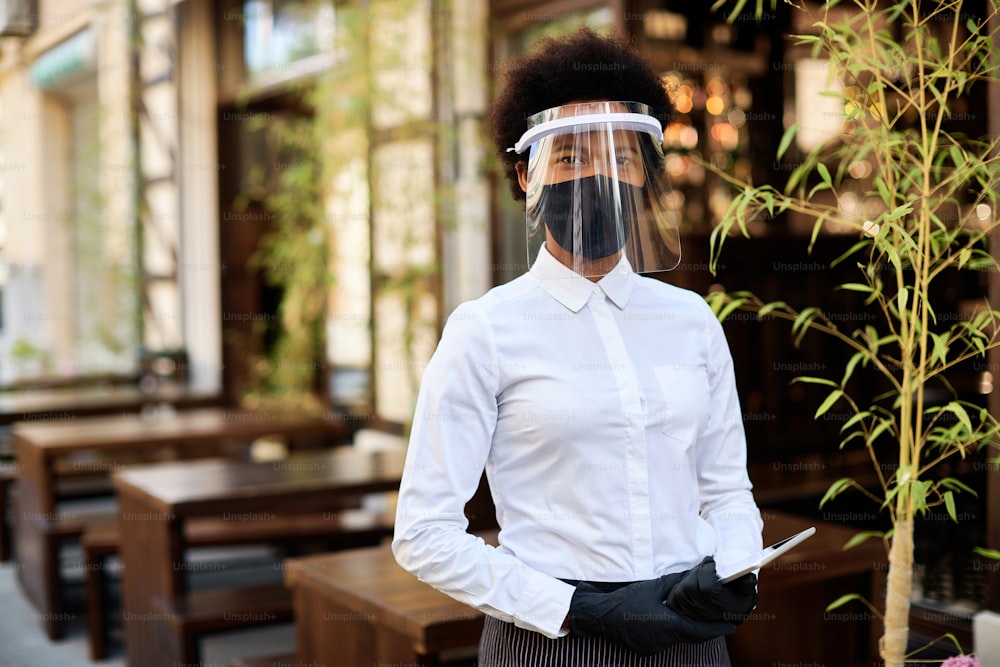 African American waitress wearing protective face mask and visor while standing at outdoor cafe and looking at camera.