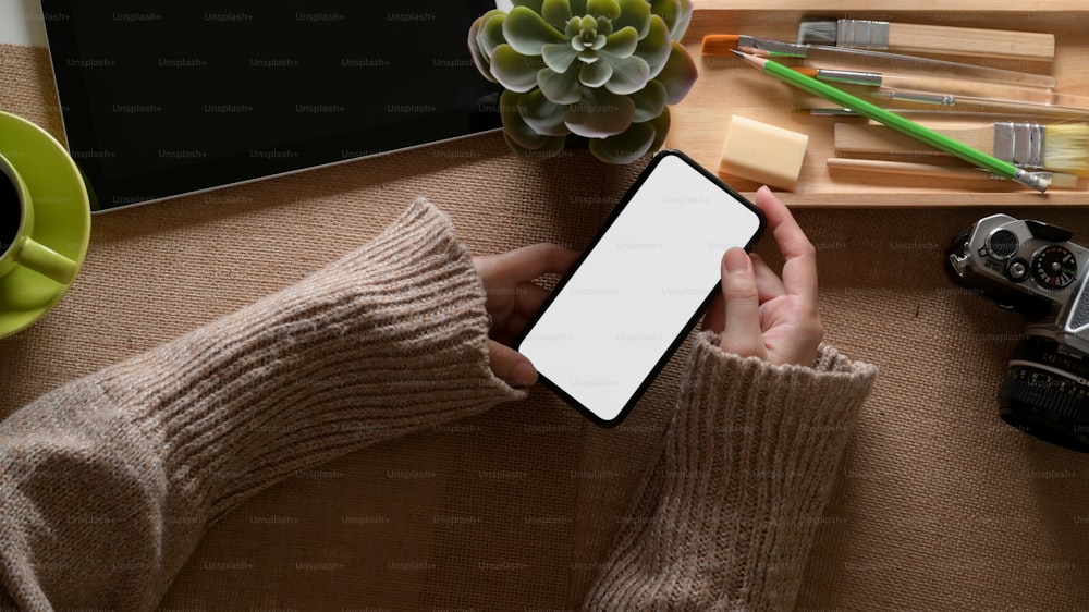 Overhead-Aufnahme einer Frau, die das Smartphone mit Clipping-Pfad auf dem Home-Office-Schreibtisch mit Zubehör hält