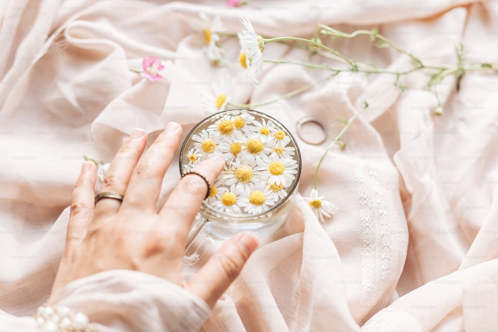 Hand mit Schmuck hält Glasbecher mit Gänseblümchen Blumen in Wasser auf dem Hintergrund von weichem beigem Stoff mit Wildblumen. Zärtliche florale Ästhetik. Kreatives Sommerimage. Bohème-Stimmung
