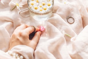 Hand holding pink wildflower on background of soft beige fabric with glass cup with daisy flowers and jewelry in sunny light. Tender floral aesthetic. Creative summer image. Bohemian mood
