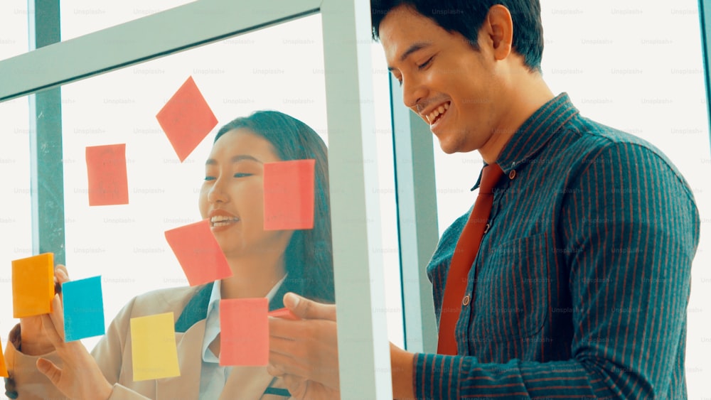 Business people work on project planning board in office and having conversation with coworker friend to analyze project development . They use sticky notes posted on glass wall to make it organized .