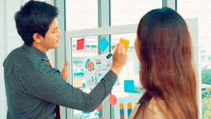 Business people work on project planning board in office and having conversation with coworker friend to analyze project development . They use sticky notes posted on glass wall to make it organized .