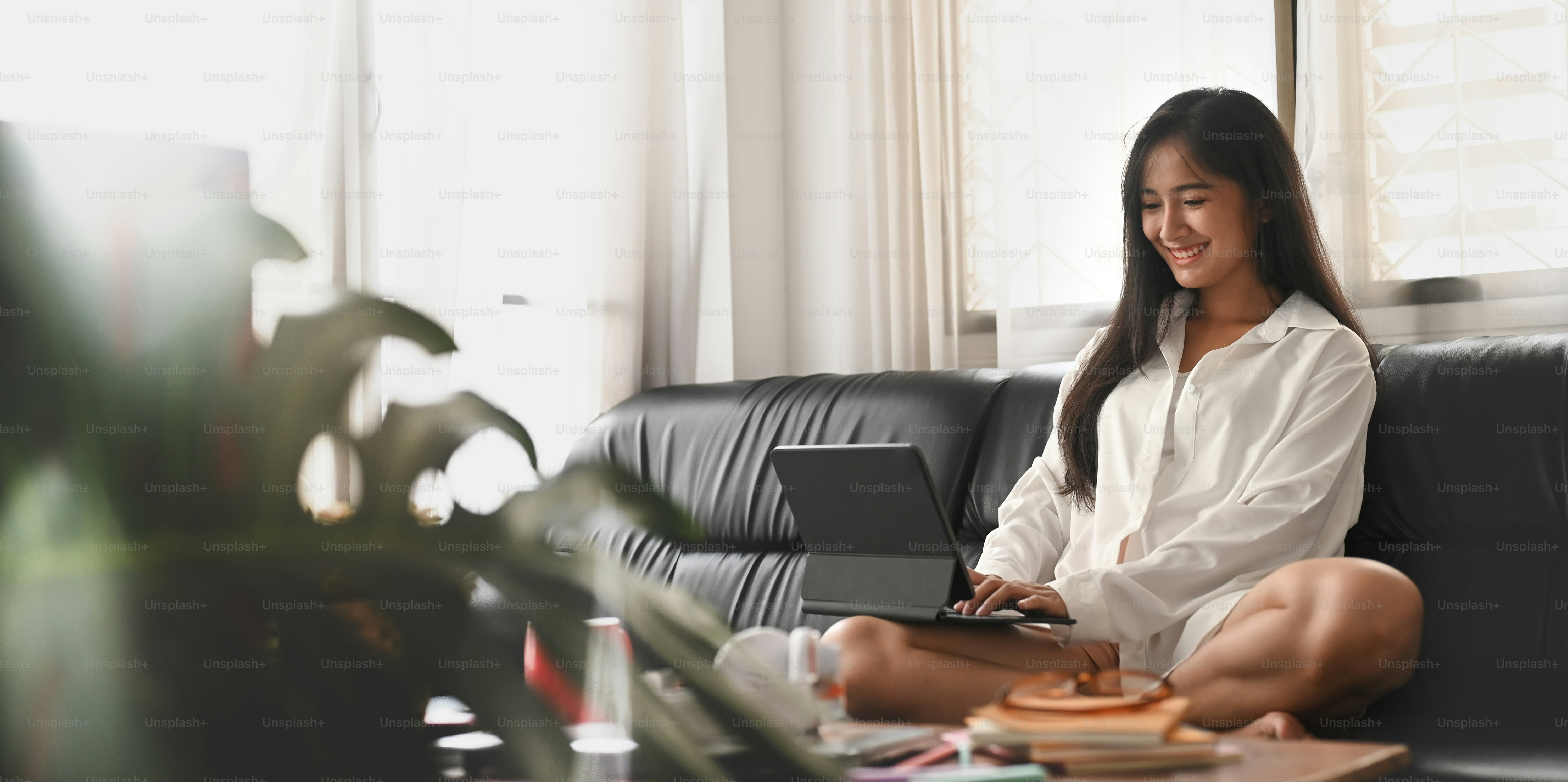A beautiful woman in a white shirt pajamas is using a computer tablet on the black sofa.