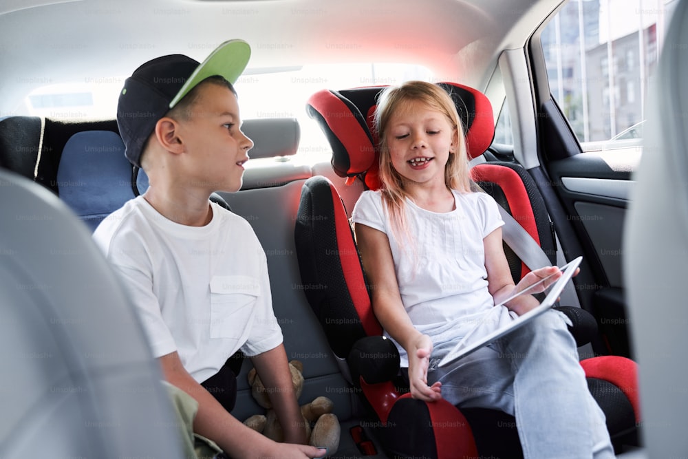 Menino e menina alegremente bonitos estão sentados em assentos de carro seguros e usando tablet digital