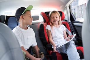 Un garçon et une fille mignons sont assis dans des sièges d’auto sûrs et utilisent une tablette numérique