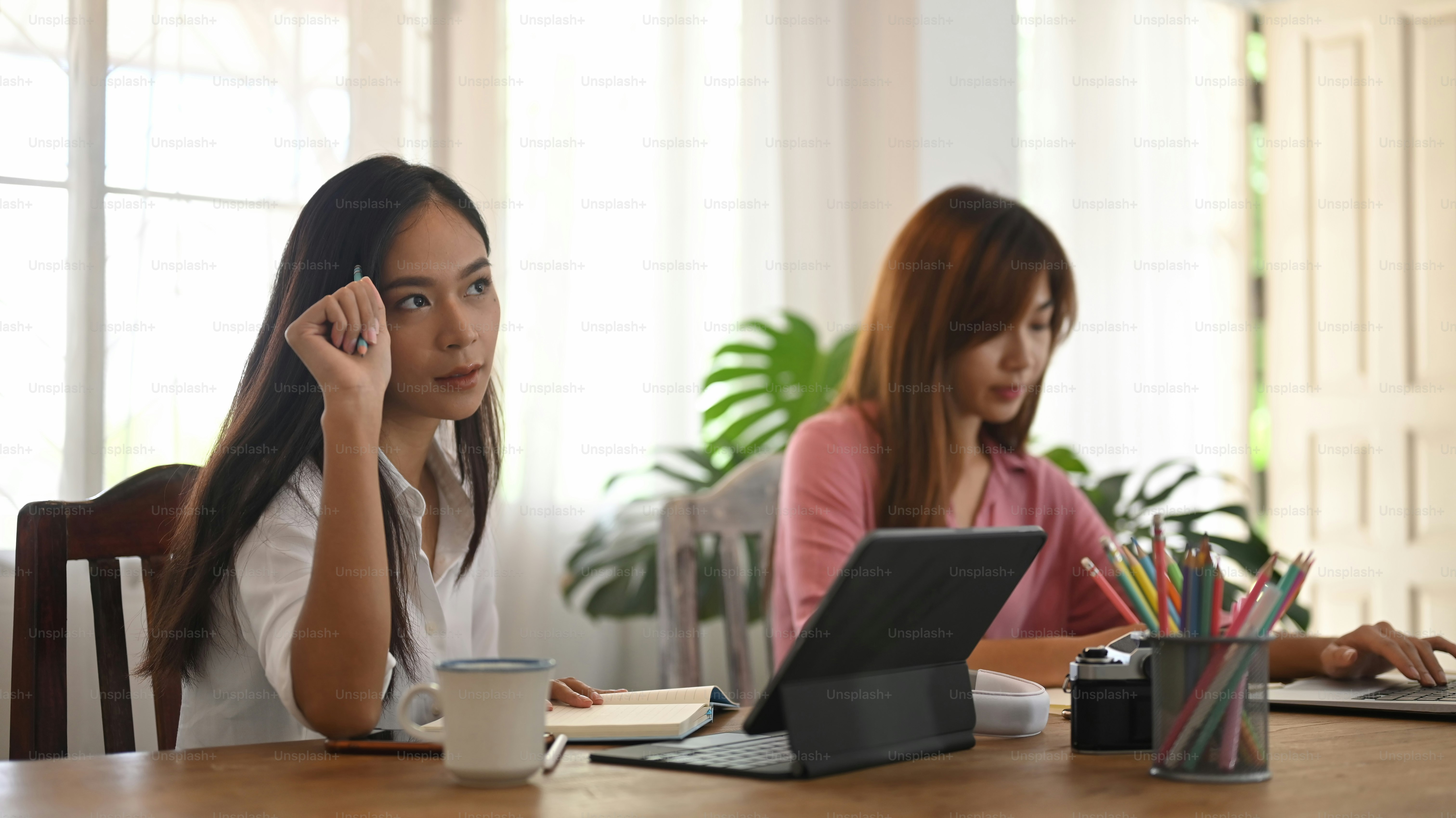 Women are using a computer tablet and laptop while sitting together at the wooden table.